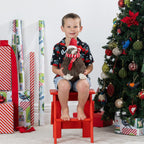 A young boy sits on a red step stool in a living room decorated for Christmas, holding a plush kiwi bird figurine with a red and white striped scarf. He is wearing a black short-sleeve holiday shirt with red and green patterns and denim shorts.