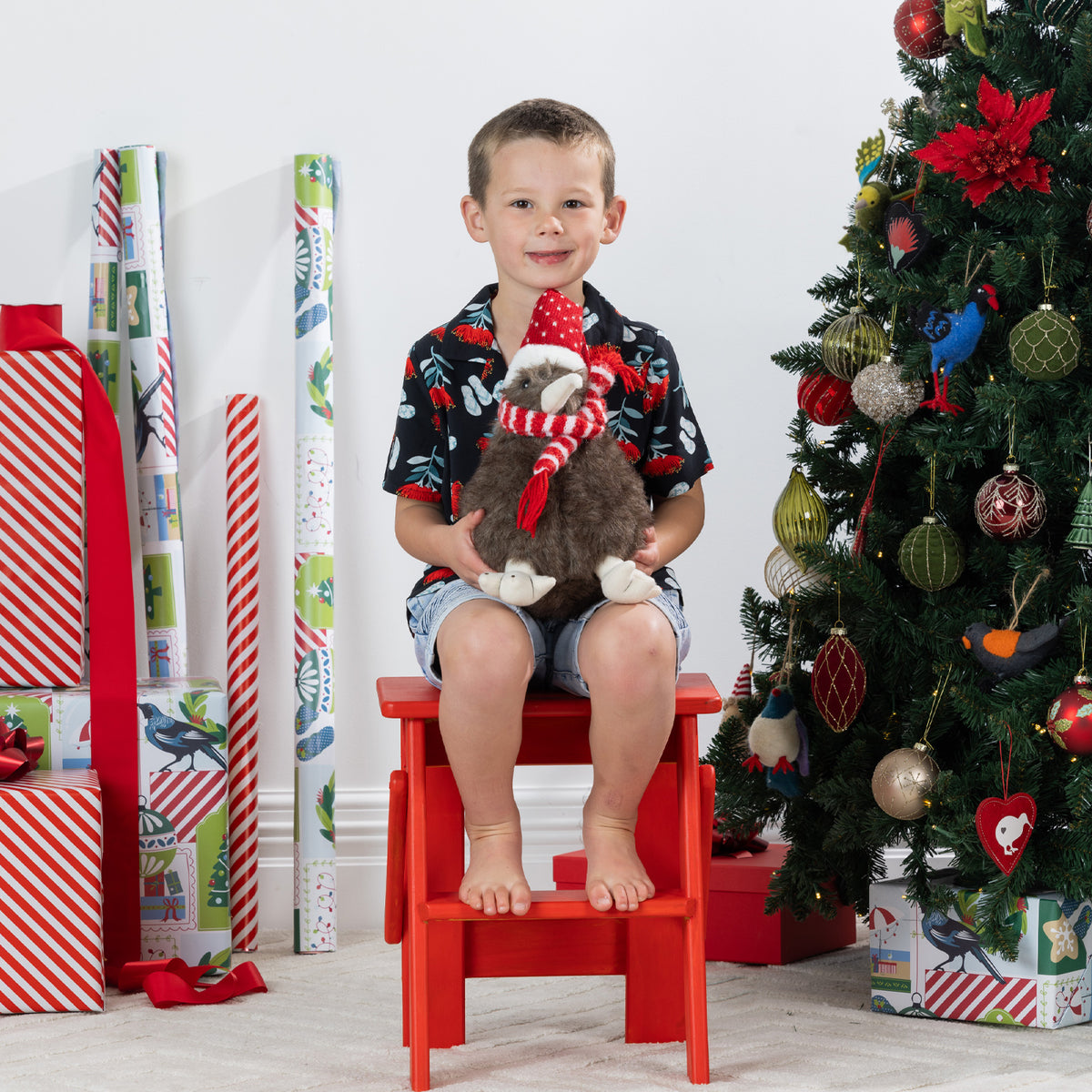 A young boy sits on a red step stool in a living room decorated for Christmas, holding a plush kiwi bird figurine with a red and white striped scarf. He is wearing a black short-sleeve holiday shirt with red and green patterns and denim shorts.