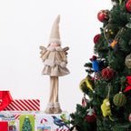 A Christmas angel figurine stands on top of wrapped presents beside a decorated Christmas tree. The figurine has a tall beige knit hat, angel wings, a cream dress, and long legs, set against a clean white background with the tree on the right.