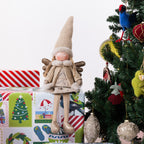 A knitted Christmas angel figurine with a tall beige hat and wings sits on top of wrapped gifts beside a decorated Christmas tree. The scene includes colorful gift boxes and holiday ornaments against a clean white background.