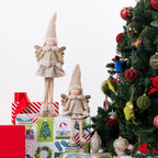Two beige angel Christmas figurines with tall pointed hats and wings, displayed on top of wrapped gifts beside a decorated Christmas tree against a white background.