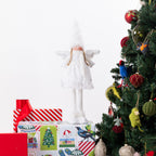 A white angel figurine with wings and a tall pointed hat stands on top of wrapped Christmas presents beside a decorated Christmas tree. The scene is photographed against a clean white background.