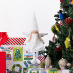 A white angel Christmas figurine with outstretched wings and a tall pointed hat, posed on top of wrapped gifts beside a decorated Christmas tree.
