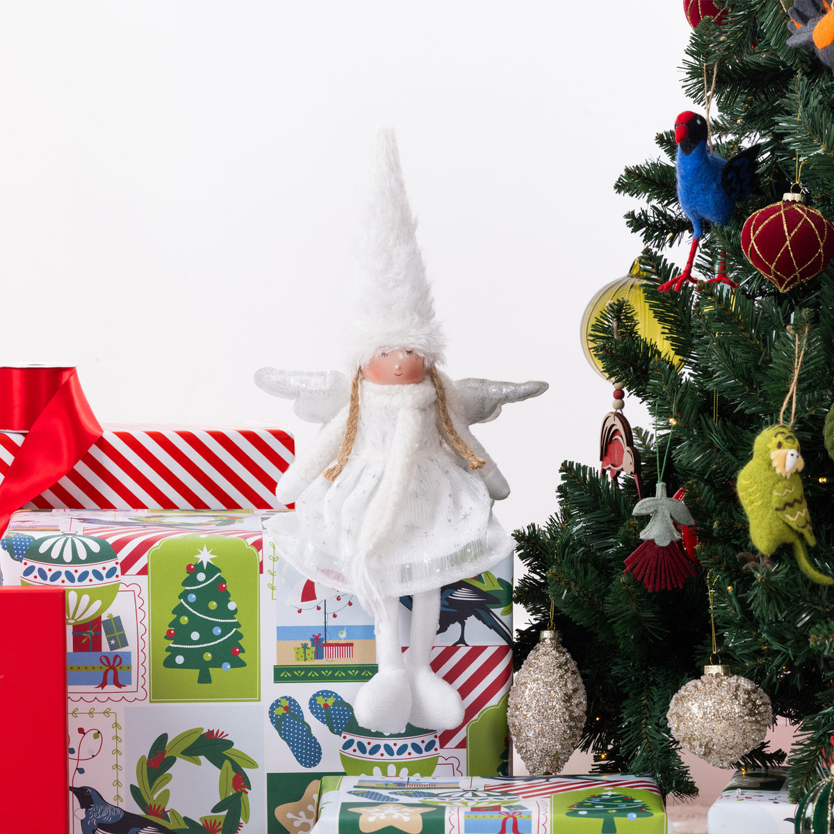 A white angel Christmas figurine with outstretched wings and a tall pointed hat, posed on top of wrapped gifts beside a decorated Christmas tree.