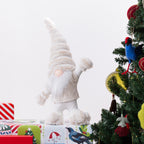 A plush Christmas gnome figurine with a tall fuzzy hat, white beard, and raised right hand, standing on wrapped gifts beside a decorated Christmas tree. 