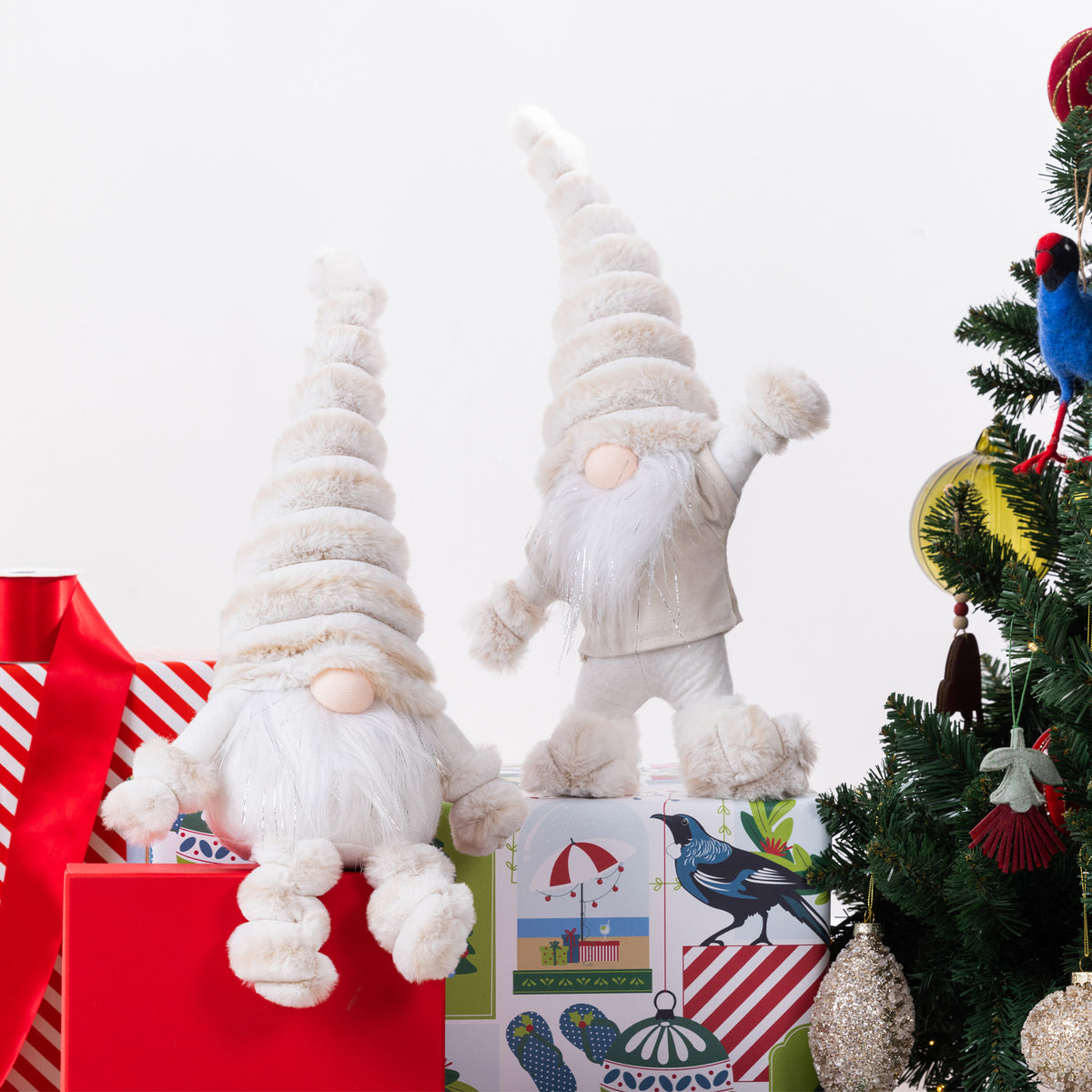 Two plush Christmas gnome figurines with tall hats and long white beards, posed among wrapped gifts and a decorated Christmas tree against a plain white background.