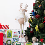 A plush Christmas reindeer figurine with long striped legs, a white fluffy body, and a striped scarf stands on wrapped gifts beside a decorated Christmas tree.