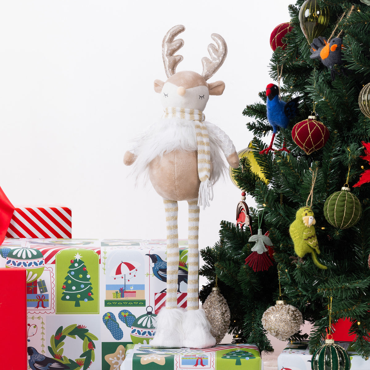 A plush Christmas reindeer figurine with long striped legs, a white fluffy body, and a striped scarf stands on wrapped gifts beside a decorated Christmas tree.