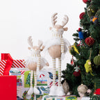 Two plush reindeer figurines with beige bodies, white fluffy trims, and striped cream-and-white scarves, posed beside a decorated Christmas tree and wrapped gifts on a white background.