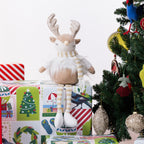 A plush Christmas reindeer figurine with long striped legs, a white fluffy body, and a striped scarf sits on wrapped gifts beside a decorated Christmas tree.