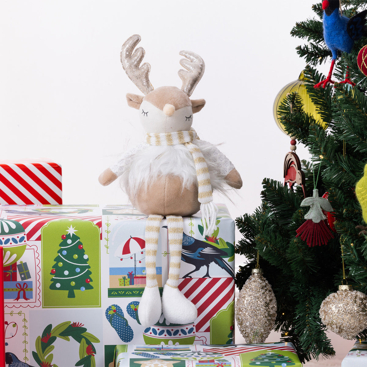 A plush Christmas reindeer figurine with long striped legs, a white fluffy body, and a striped scarf sits on wrapped gifts beside a decorated Christmas tree.