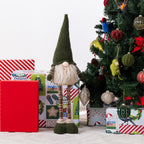 A Christmas gnome figurines stands in front of wrapped gifts and a decorated Christmas tree. The gnome has a tall dark green pointed hat, a white beard, striped brown legs, and dark green shoes.