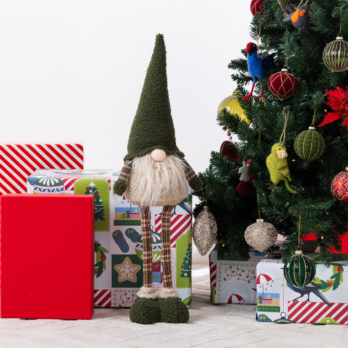 A Christmas gnome figurines stands in front of wrapped gifts and a decorated Christmas tree. The gnome has a tall dark green pointed hat, a white beard, striped brown legs, and dark green shoes.