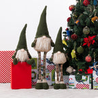 Three knitted Christmas gnome figurines with tall dark green pointed hats, white beards, and plaid legs, arranged beside a decorated Christmas tree and wrapped presents on a white studio background.