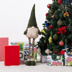 A Christmas gnome figurine stands in front of wrapped gifts and a decorated Christmas tree. The gnome has a tall dark green pointed hat, a white beard, striped brown legs, and dark green shoes.