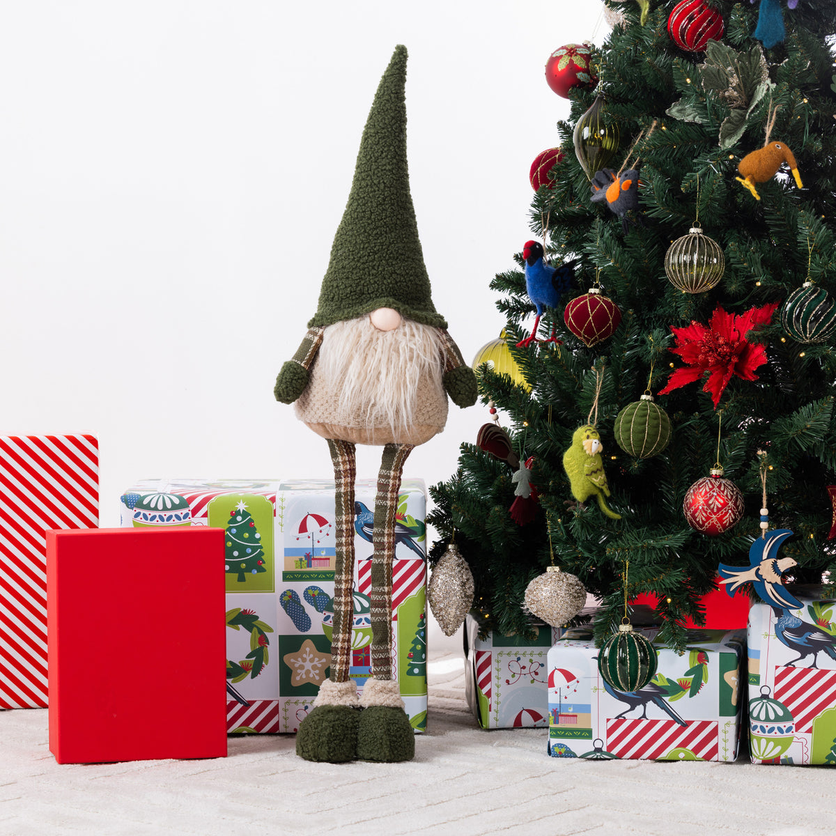 A Christmas gnome figurine stands in front of wrapped gifts and a decorated Christmas tree. The gnome has a tall dark green pointed hat, a white beard, striped brown legs, and dark green shoes.