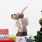 A plush Christmas moose figurine with a long hat, brown antlers and hooves, and fluffy beige fur, posed on top of wrapped gifts against a white background with a Christmas tree edge visible on the right.
