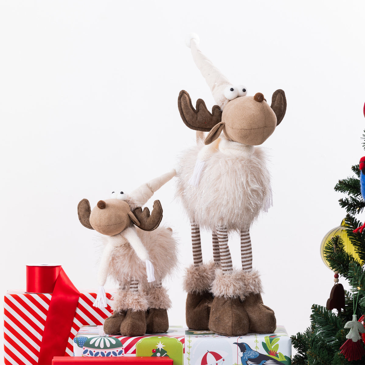 Two plush moose figurines with fluffy cream fur, brown antlers and hooves, and striped legs, posed on top of wrapped presents beside a decorated Christmas tree against a white background.