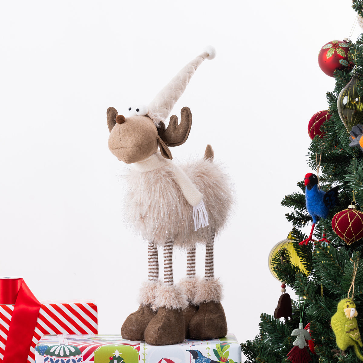 A plush moose Christmas figurine with a cream faux-fur body, brown antlers and hooves, and a long cream hat, standing on wrapped gifts beside a decorated Christmas tree.