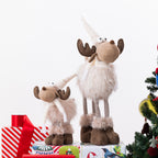Two plush moose figurines with fluffy cream fur, brown antlers and hooves, and striped legs, posed on top of wrapped presents beside a decorated Christmas tree against a white background.