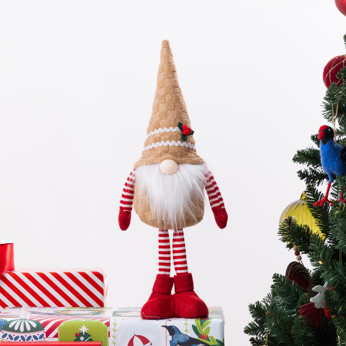 A Christmas gnome plush figurine with a tall gingerbread hat, white beard, red-and-white striped arms and legs, and red shoes, posed on top of wrapped gifts beside a decorated Christmas tree.