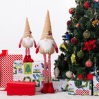Two Christmas gnome figurines standing beside wrapped gifts and a decorated Christmas tree. The gnomes have gingerbread pointed hats, white beards, red-and-white striped legs, and red boots.