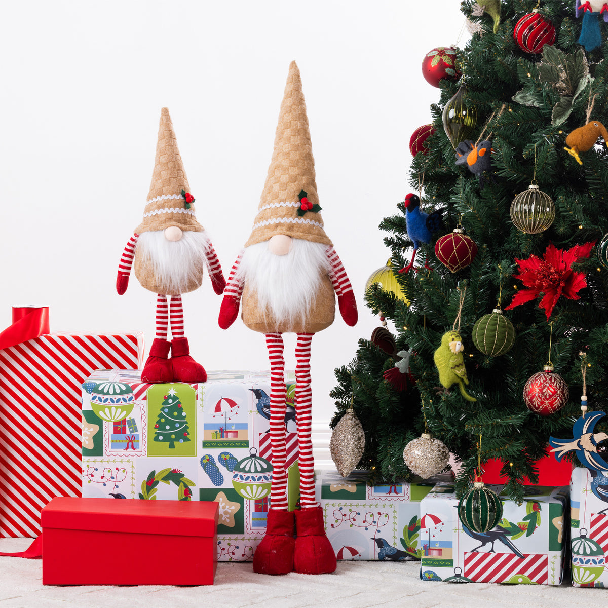 Two Christmas gnome figurines standing beside wrapped gifts and a decorated Christmas tree. The gnomes have gingerbread pointed hats, white beards, red-and-white striped legs, and red boots.