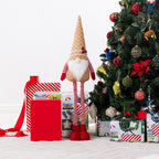 A Christmas gnome plush figurine stands under a decorated Christmas tree beside wrapped gifts and a red gift box on a light floor against a white wall.