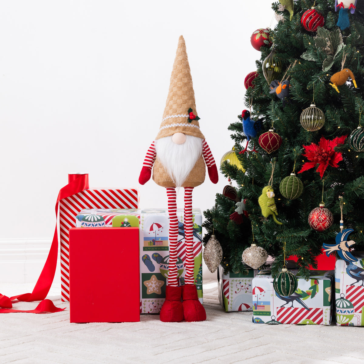 A Christmas gnome plush figurine stands under a decorated Christmas tree beside wrapped gifts and a red gift box on a light floor against a white wall.