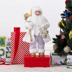 Christmas Wishes White & Gold Santa 60cm dressed in a white and gold festive outfit, holding silver baubles and a sack of decorations beside a Christmas tree and wrapped presents.