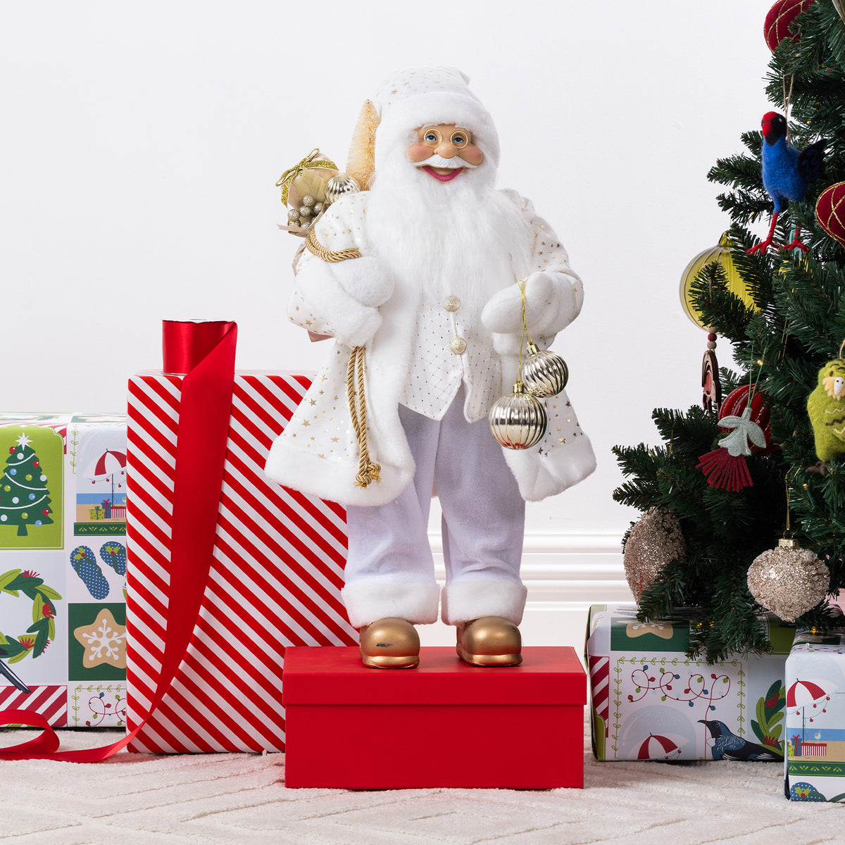Christmas Wishes White & Gold Santa 60cm dressed in a white and gold festive outfit, holding silver baubles and a sack of decorations beside a Christmas tree and wrapped presents.