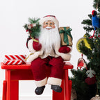 Christmas Wishes Red Traditional Sitting Santa 40cm dressed in a red festive outfit with white fur trim, holding a gold gift and a sack of Christmas greenery while sitting beside a decorated Christmas tree.