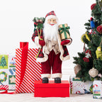 Christmas Wishes Red Traditional Santa 60cm dressed in a red tartan coat with white fur trim, holding a gold gift and a sack of Christmas greenery beside a decorated Christmas tree.