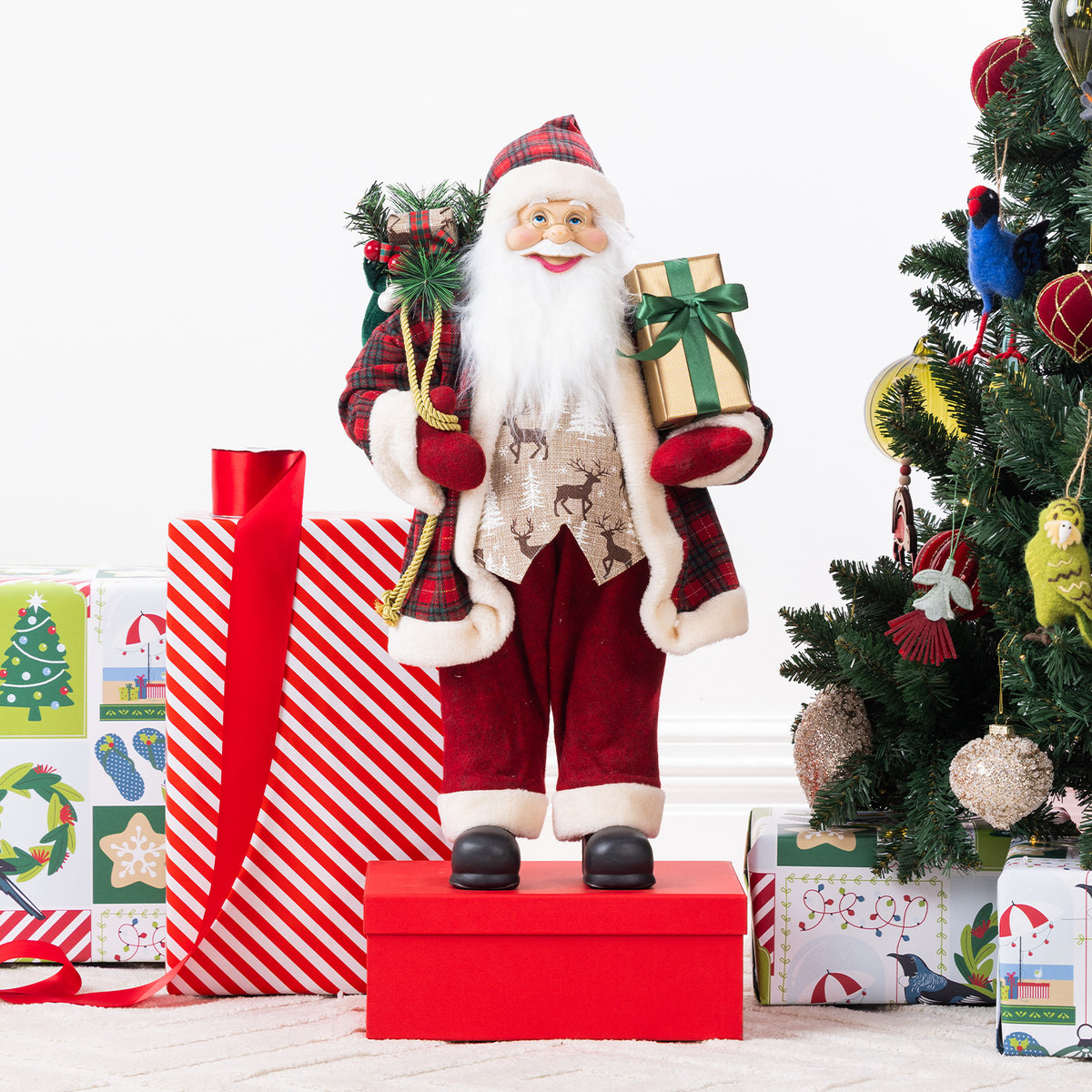 Christmas Wishes Red Traditional Santa 60cm dressed in a red tartan coat with white fur trim, holding a gold gift and a sack of Christmas greenery beside a decorated Christmas tree.