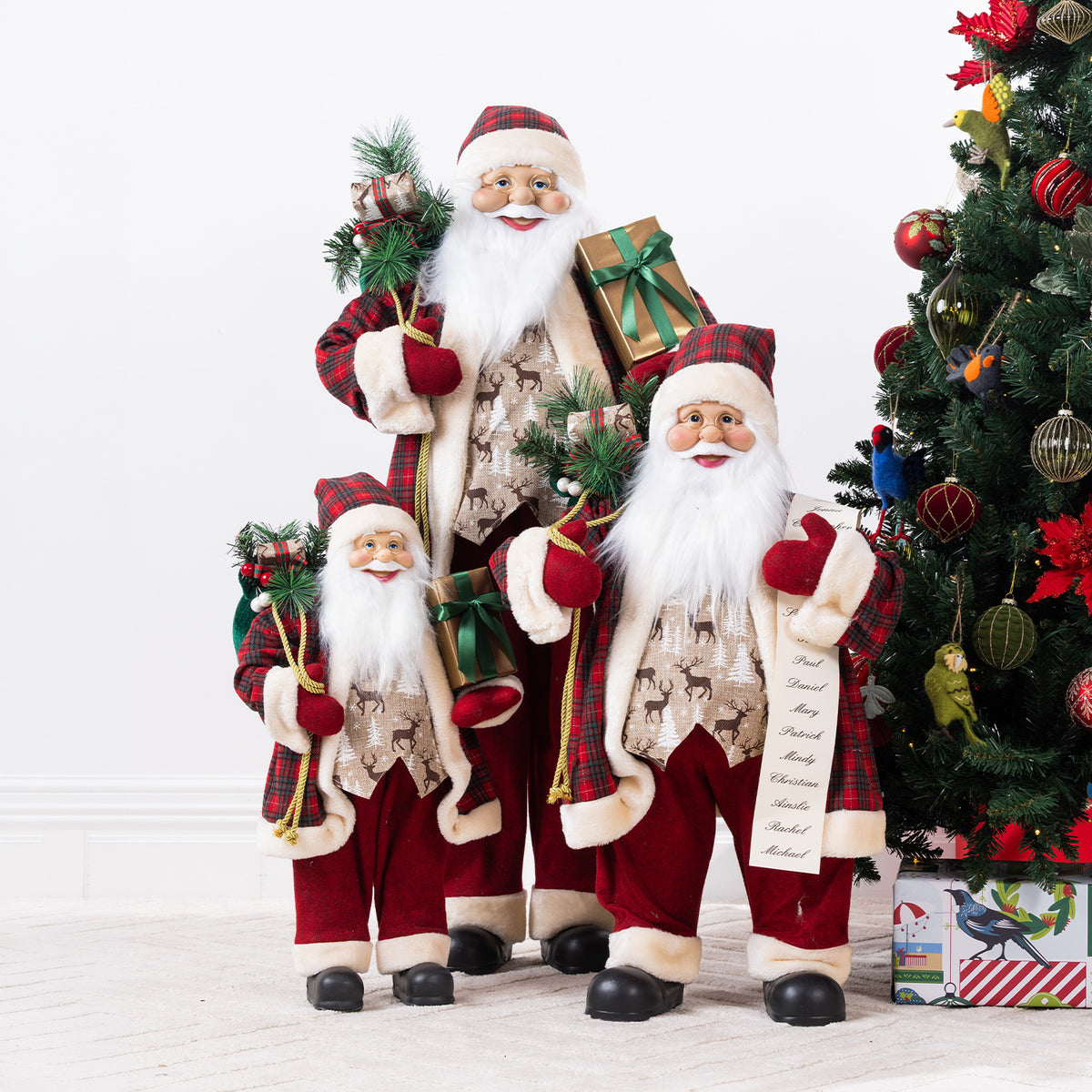 Three Christmas Wishes Red Traditional Santa figures in different sizes, wearing matching red tartan outfits and displayed beside a decorated Christmas tree.