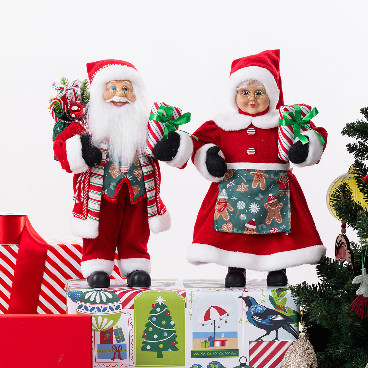 Christmas Wishes Gingerbread Mr and Mrs Claus figures dressed in festive red outfits, holding wrapped presents and displayed beside a decorated Christmas tree.