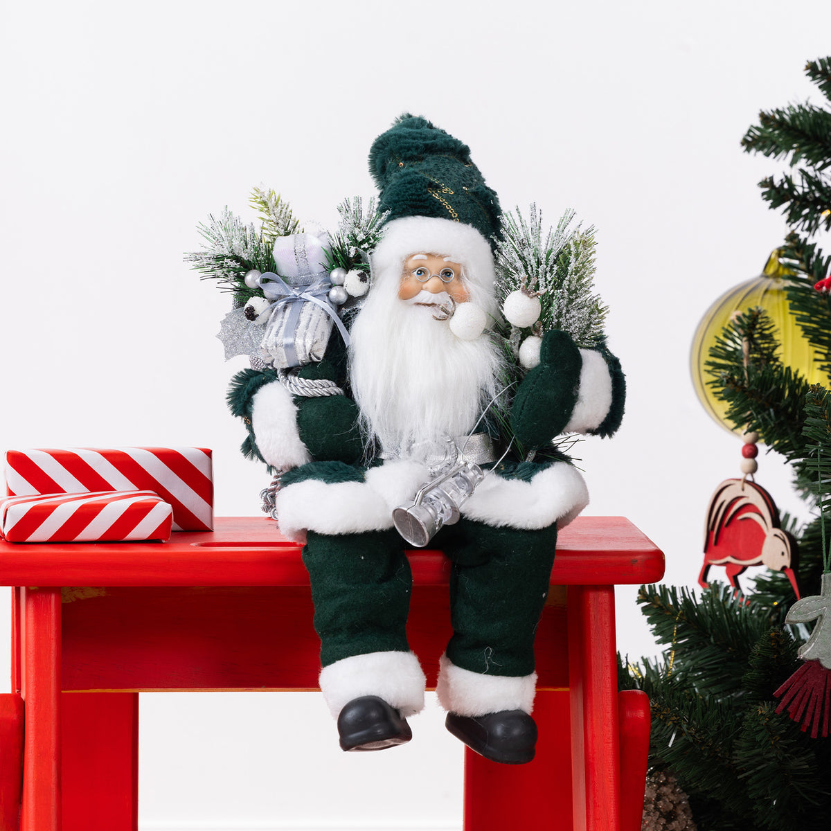 Christmas Wishes Green Sitting Santa 30cm dressed in a deep green and white outfit, holding silver festive decorations and surrounded by frosted greenery.
