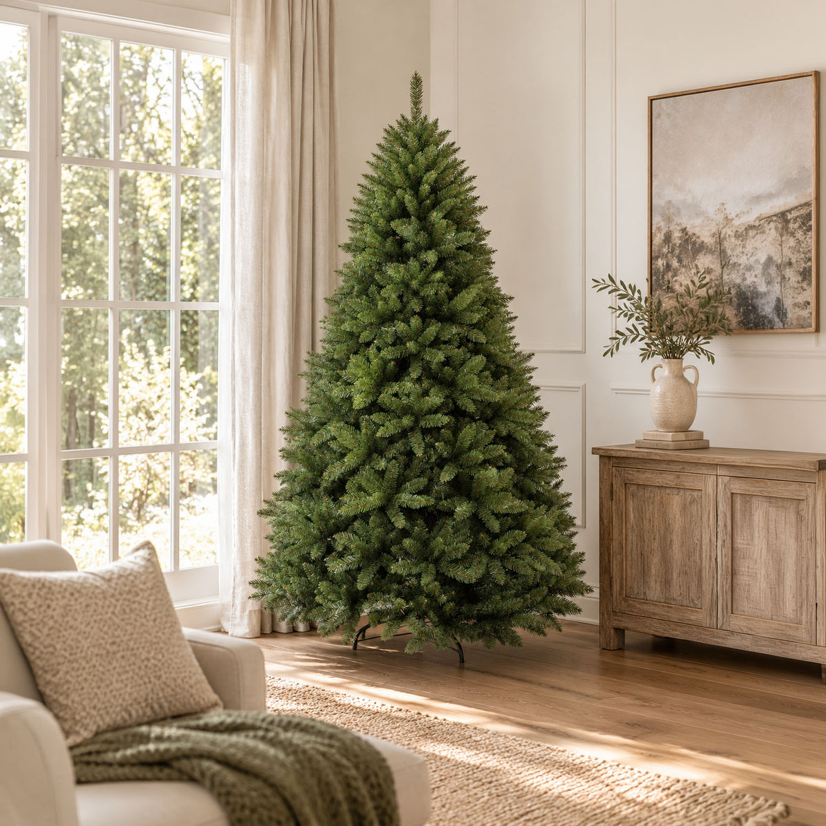 A green artificial Christmas tree stands in a bright living room corner next to a large window, with a beige armchair, wood cabinet, and framed wall art in the background.