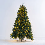 A pre-lit artificial Christmas tree with green needles and warm yellow lights, standing on a black metal stand against a plain white wall and light floor.