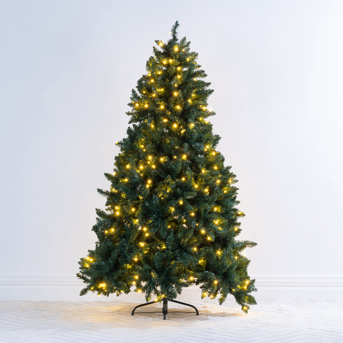 A pre-lit artificial Christmas tree with green needles and warm yellow lights, standing on a black metal stand against a plain white wall and light floor.