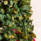 A close-up of a green artificial Christmas tree with warm yellow lights glowing through the branches, with a red ribbon or skirt visible at the base against a light indoor wall.