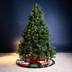 A decorated artificial Christmas tree with warm white string lights stands on a black circular toy train track. Under the tree are several small holiday-themed gift boxes, and a red Santa-themed base sits at the bottom.
