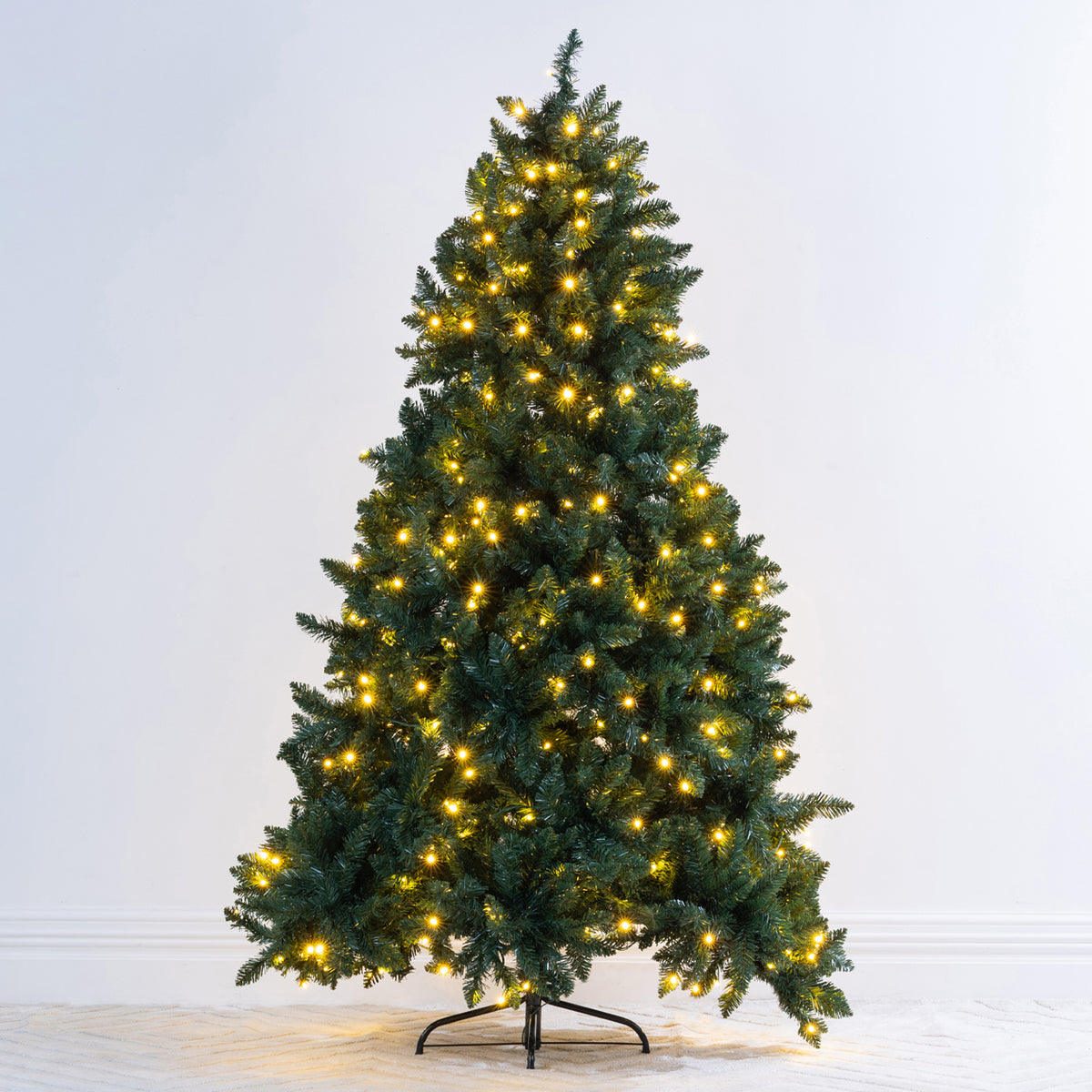 A pre-lit artificial Christmas tree with green needles and warm yellow lights, standing on a black metal stand against a plain white wall.
