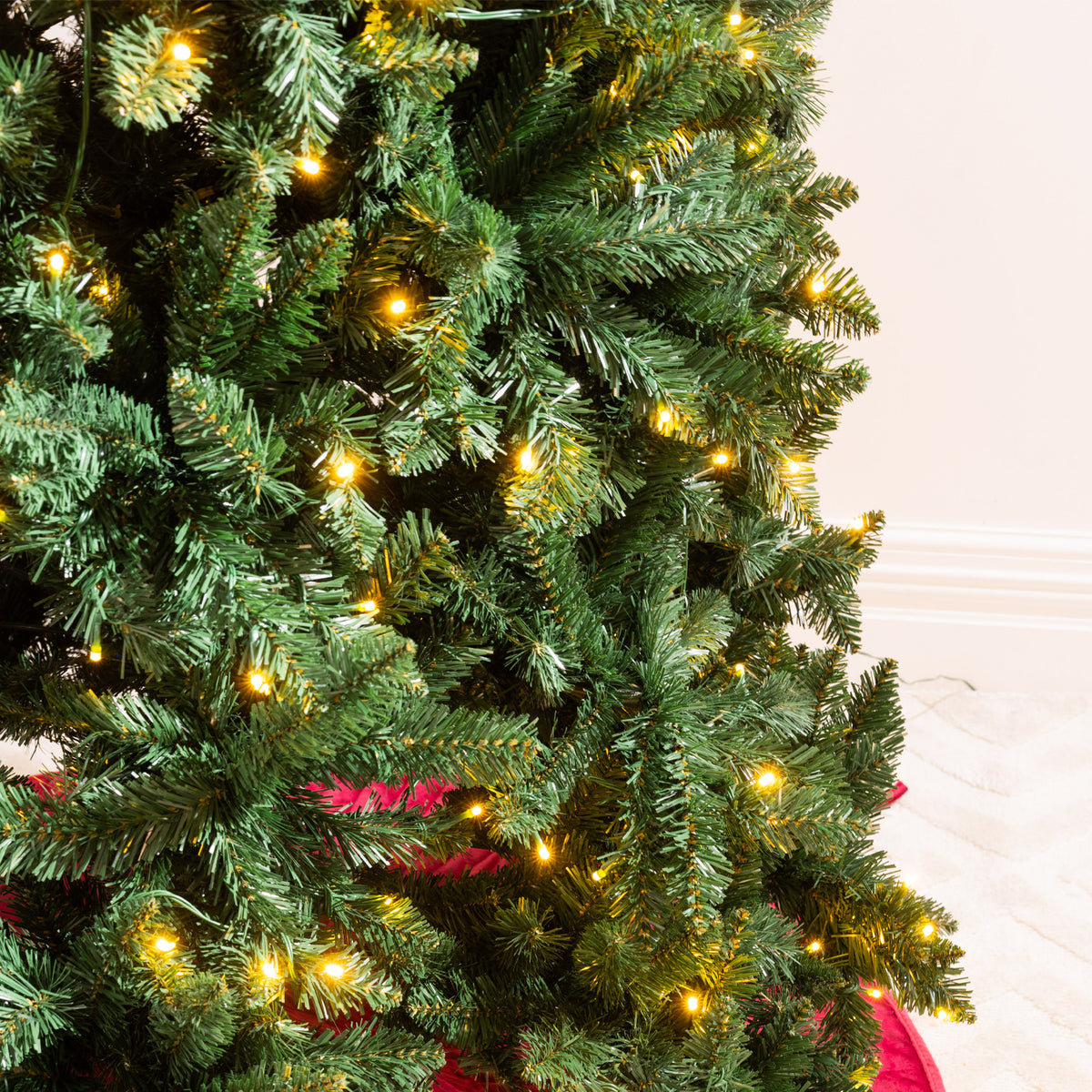 A close-up of a green artificial Christmas tree with warm yellow lights glowing through the branches, with a red ribbon or skirt visible at the base against a light indoor wall.