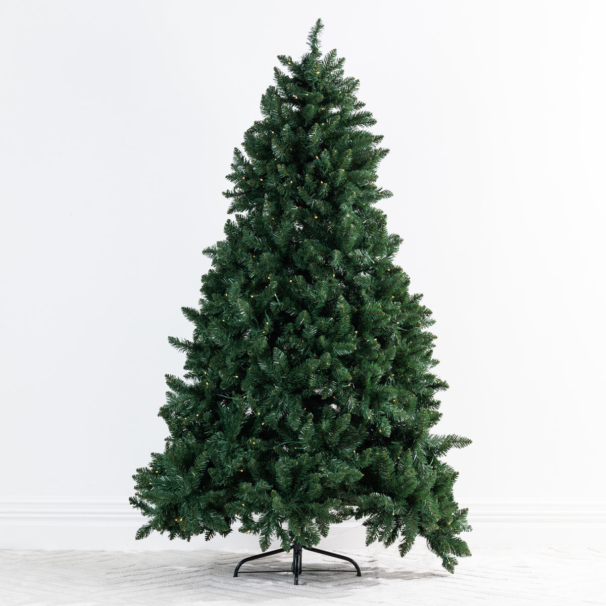 A pre-lit artificial Christmas tree with dense dark green needles on a black metal stand, photographed against a clean white studio background.
