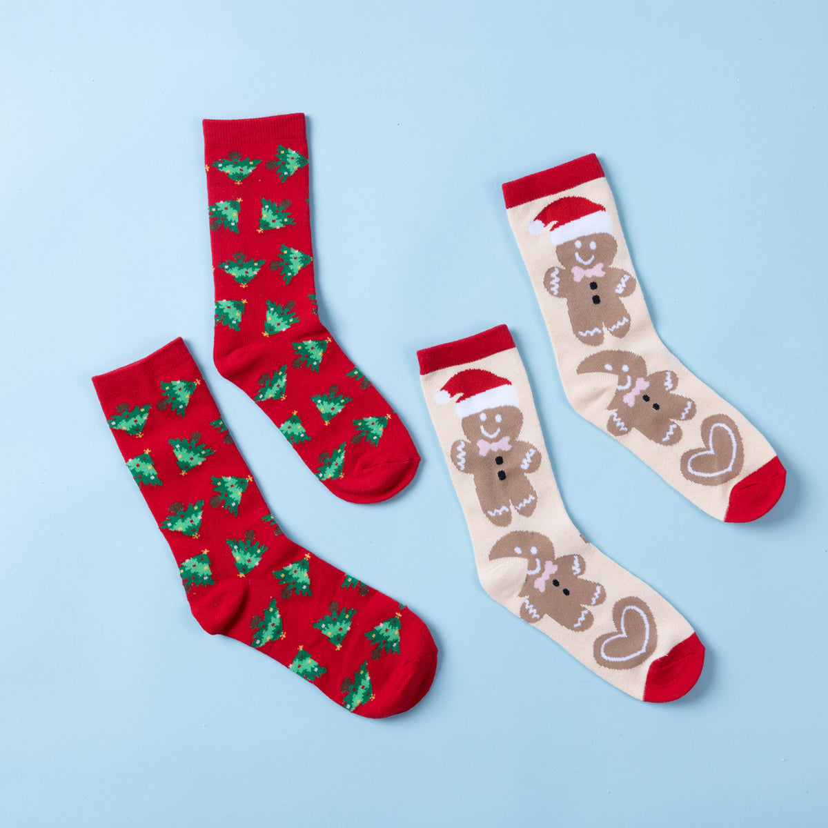 Two pairs of Christmas socks featuring red Christmas tree prints and a cream gingerbread design with Santa hats, hearts and red accents, displayed on a light blue background.