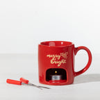 Red Christmas fondue mug with a lit tealight candle underneath and two fondue forks displayed alongside.