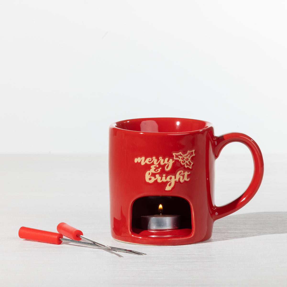 Red Christmas fondue mug with a lit tealight candle underneath and two fondue forks displayed alongside.