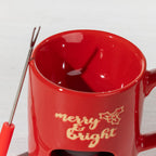 Close-up of a red Christmas fondue mug with a chocolate melting bowl, two fondue forks and gold “Merry & Bright” detailing.