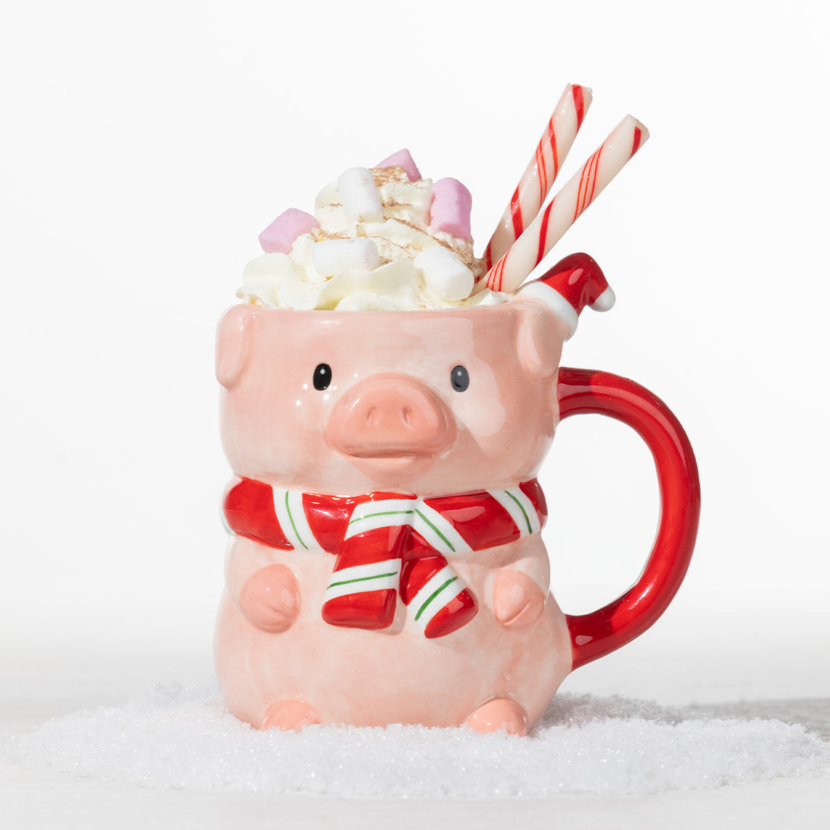 Christmas pig mug with a red and white striped scarf, filled with whipped cream, marshmallows and candy cane sticks in a snowy festive setting.
